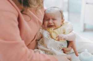 A baby is being held by a person with long hair wearing a peach top. The baby is crying and is wearing a floral vest and yellow bib.